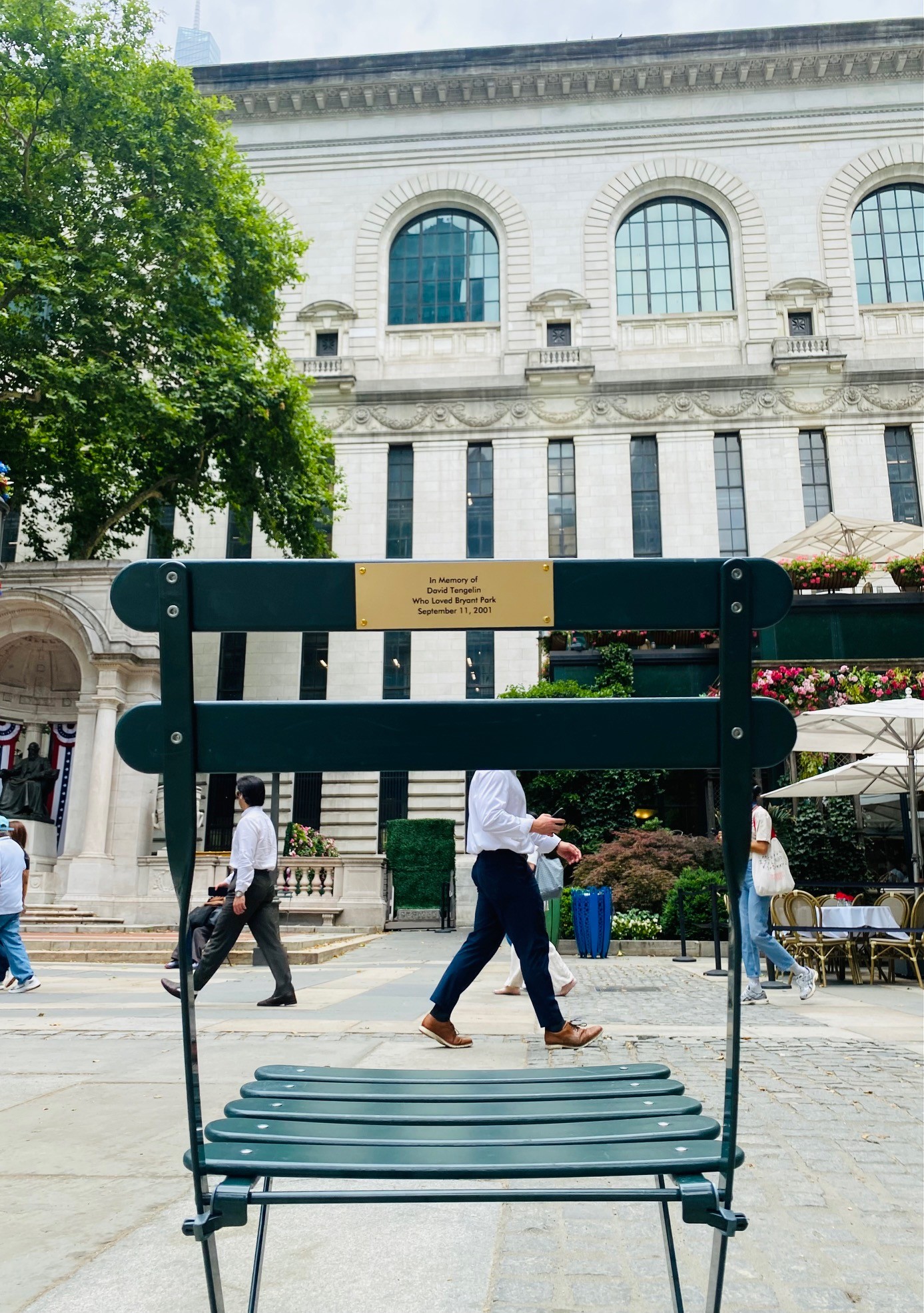 Green Bryant Park memorial chair viewed from behind with commemorative plaque visible, facing the New York Public Library. Café umbrellas, summer trees, and pedestrians moving through the park appear in the background.